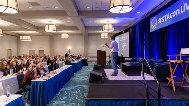 Wide view of the STACon Live ballroom with a speaker presenting on stage.